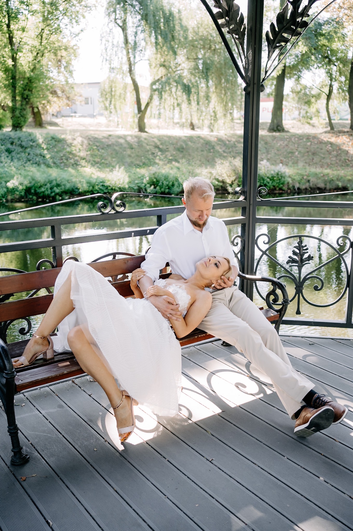 Para młoda odpoczywa na ławce w altanie nad wodą, panna młoda w sukni z koronkową górą i złotych sandałach leży na kolanach pana młodego.
