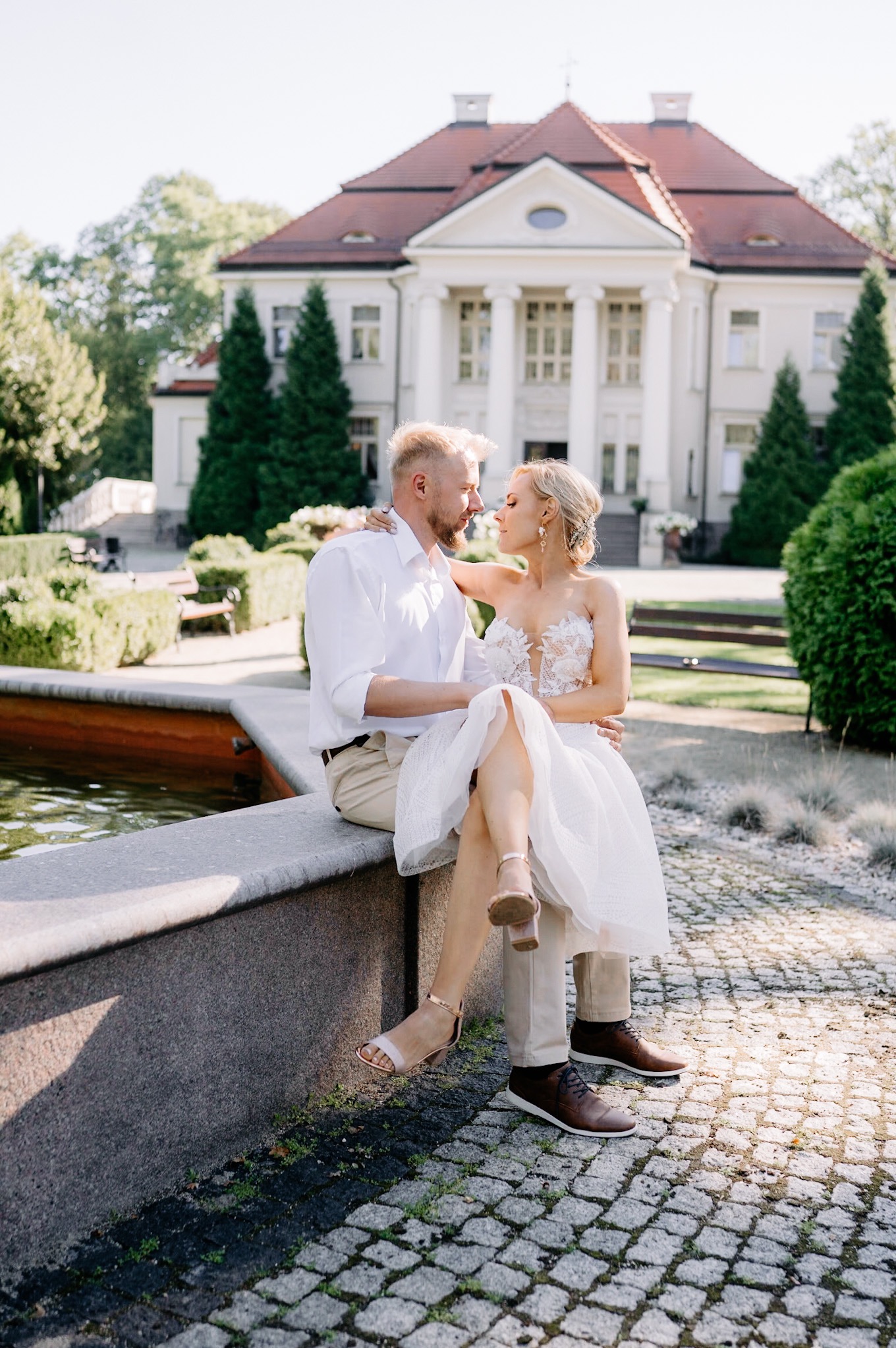 Para młoda siedzi na kamiennym murku fontanny, na tle klasycystycznego pałacu i zieleni, panna młoda w krótkiej sukience z koronkową górą i złotych sandałkach.