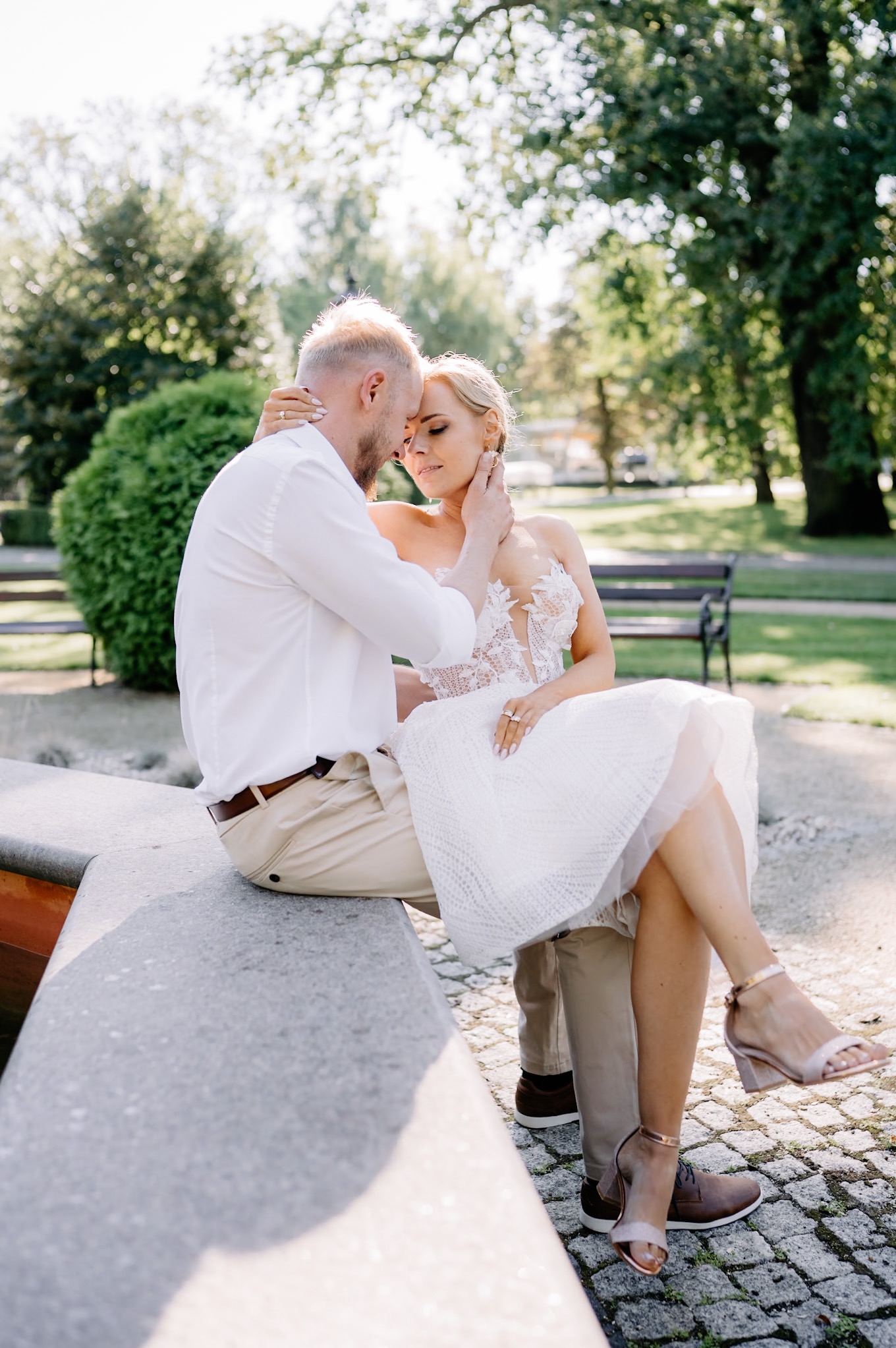Para młoda w objęciach siedzi na kamiennym murku fontanny w parku, panna młoda w krótkiej koronkowej sukience i sandałach na obcasie, pan młody w białej koszuli i beżowych spodniach.
