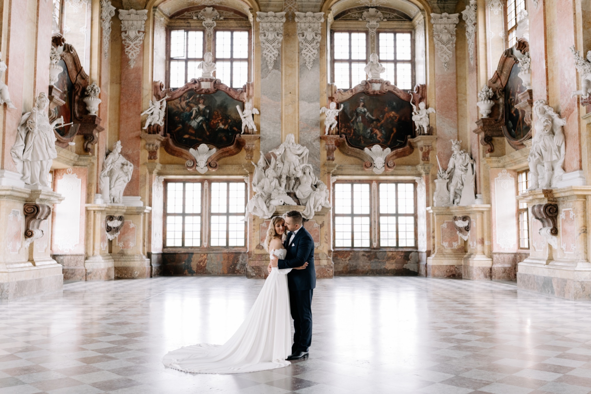 Para młoda w eleganckim uścisku na tle barokowego wnętrza z marmurową podłogą, rzeźbami i obrazami w bogato zdobionych ramach.