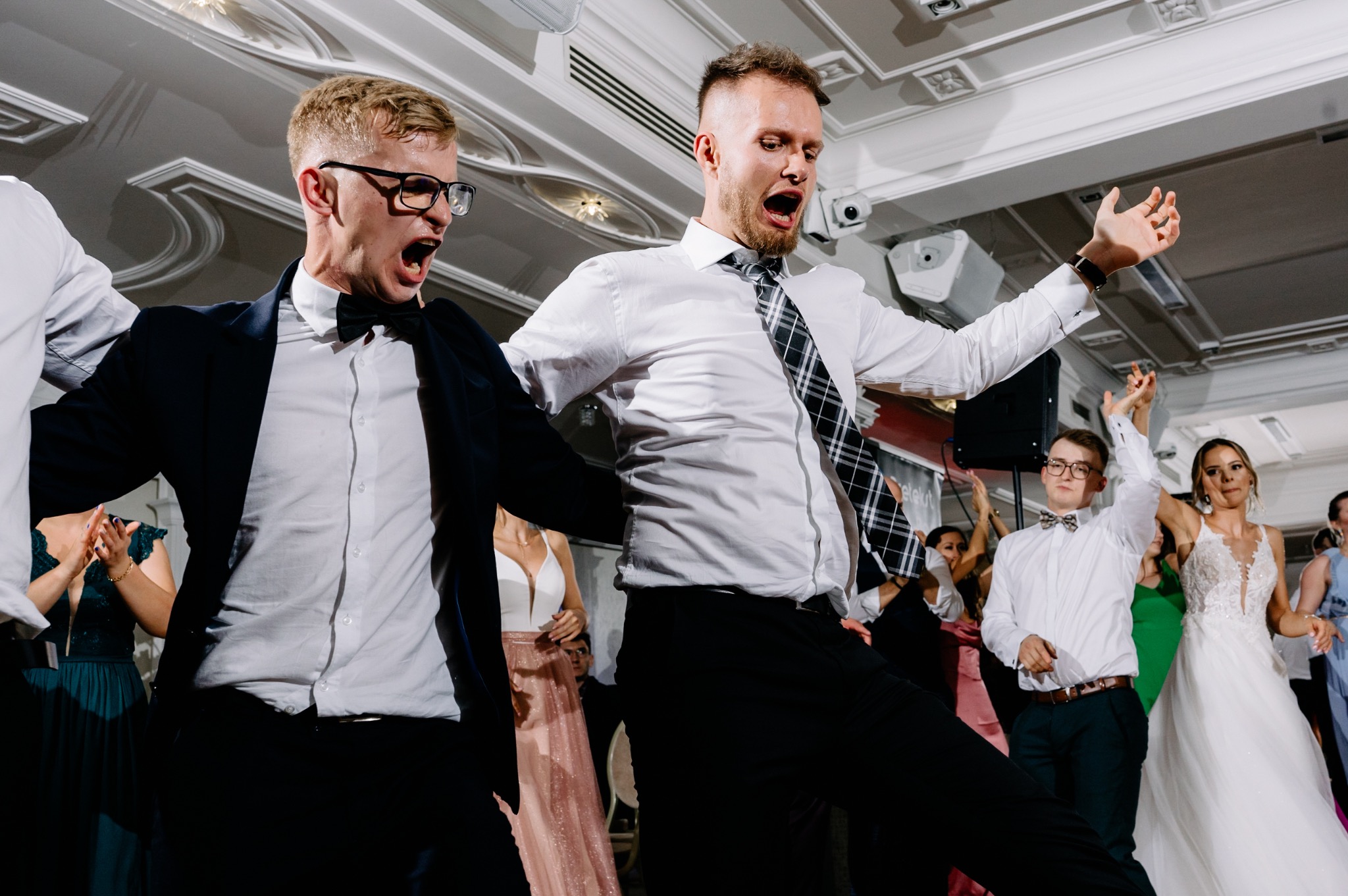 Rozbawieni goście weselni tańczą i śpiewają z otwartymi ustami, widoczna para młoda w tle, oświetlenie parkietu.