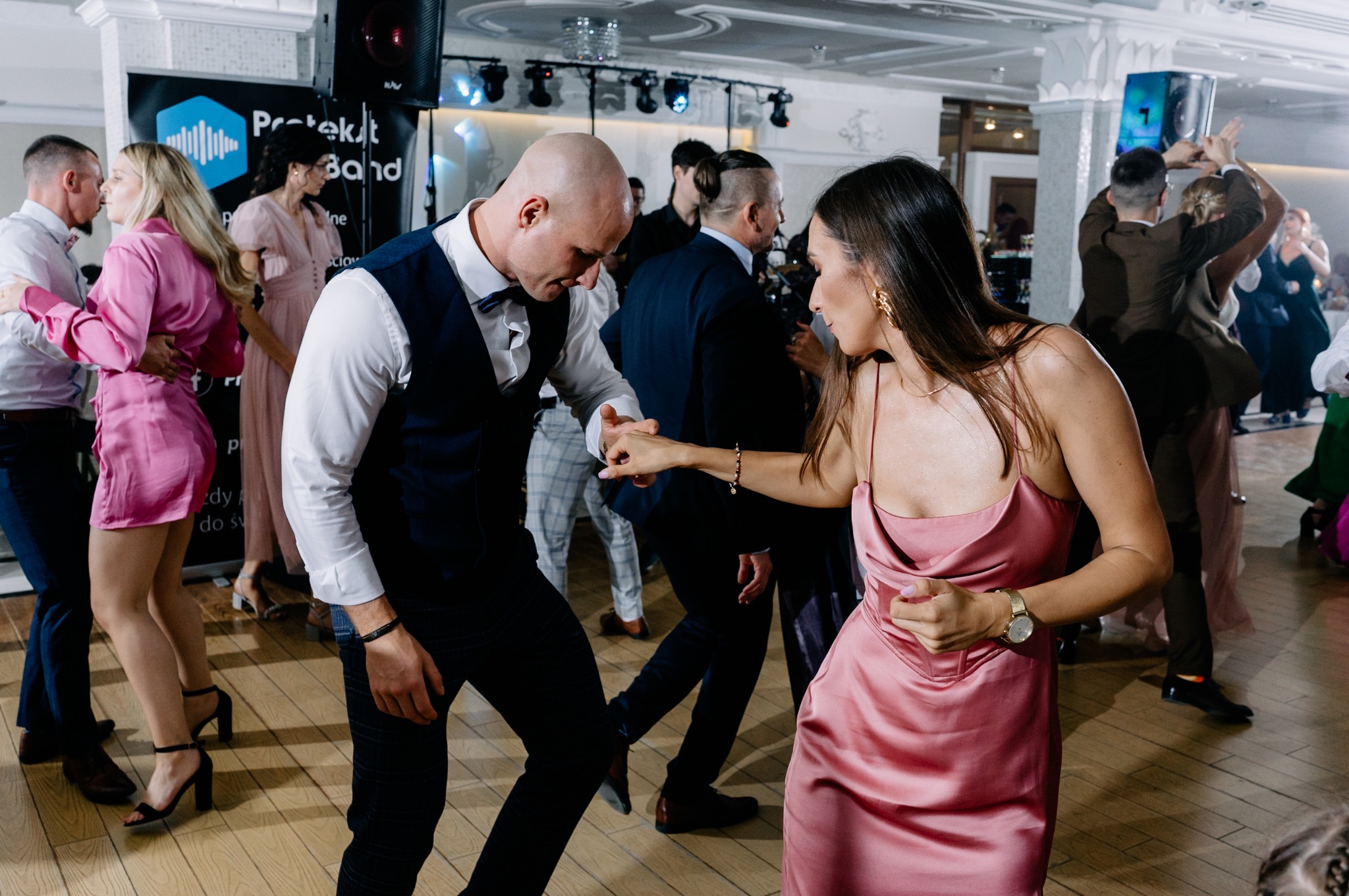 Para w różowych strojach tańczy na parkiecie podczas imprezy, w tle widoczni inni goście i sprzęt nagłośnieniowy.