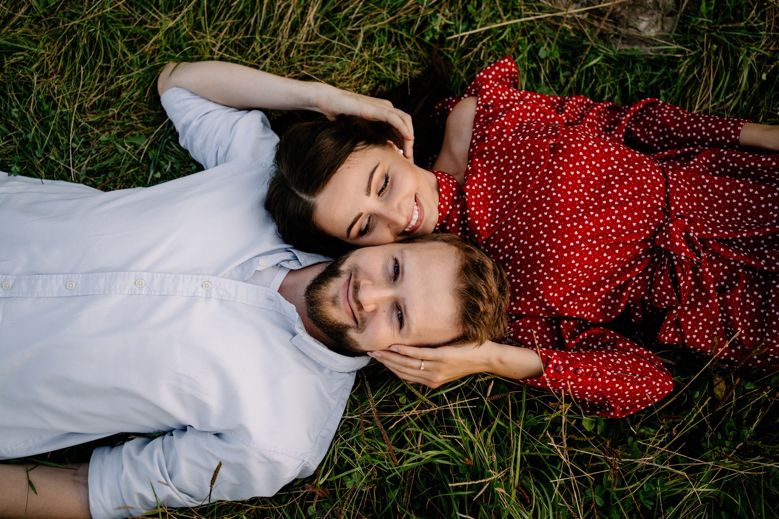 Uśmiechnięta para leży na trawie: kobieta w czerwonej sukience w białe serduszka i mężczyzna w błękitnej koszuli, obejmują się i patrzą w górę.