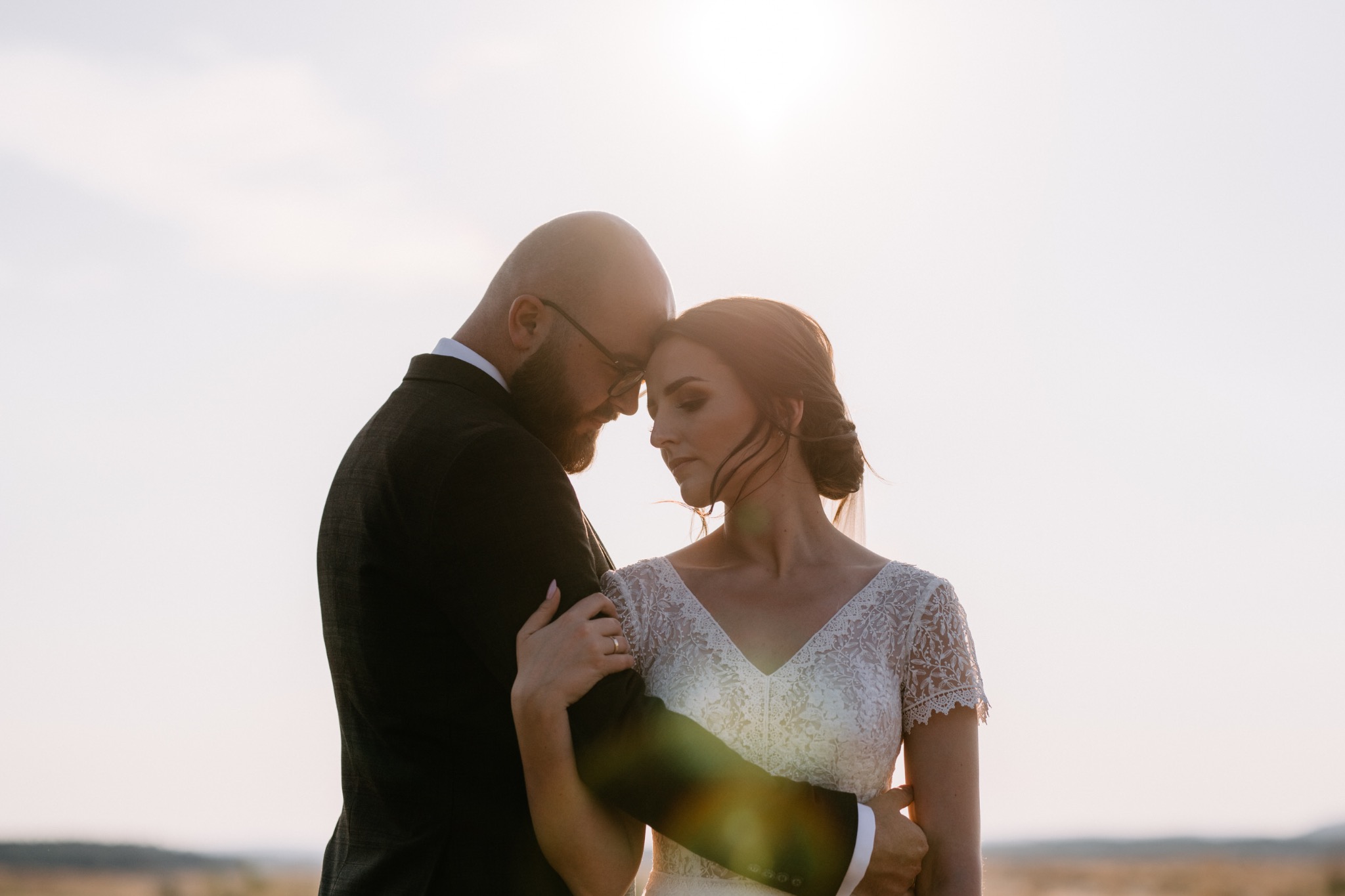 Para młoda w objęciach na tle rozświetlonego nieba, panna młoda w koronkowej sukni z krótkim rękawem, pan młody w ciemnym garniturze i okularach.