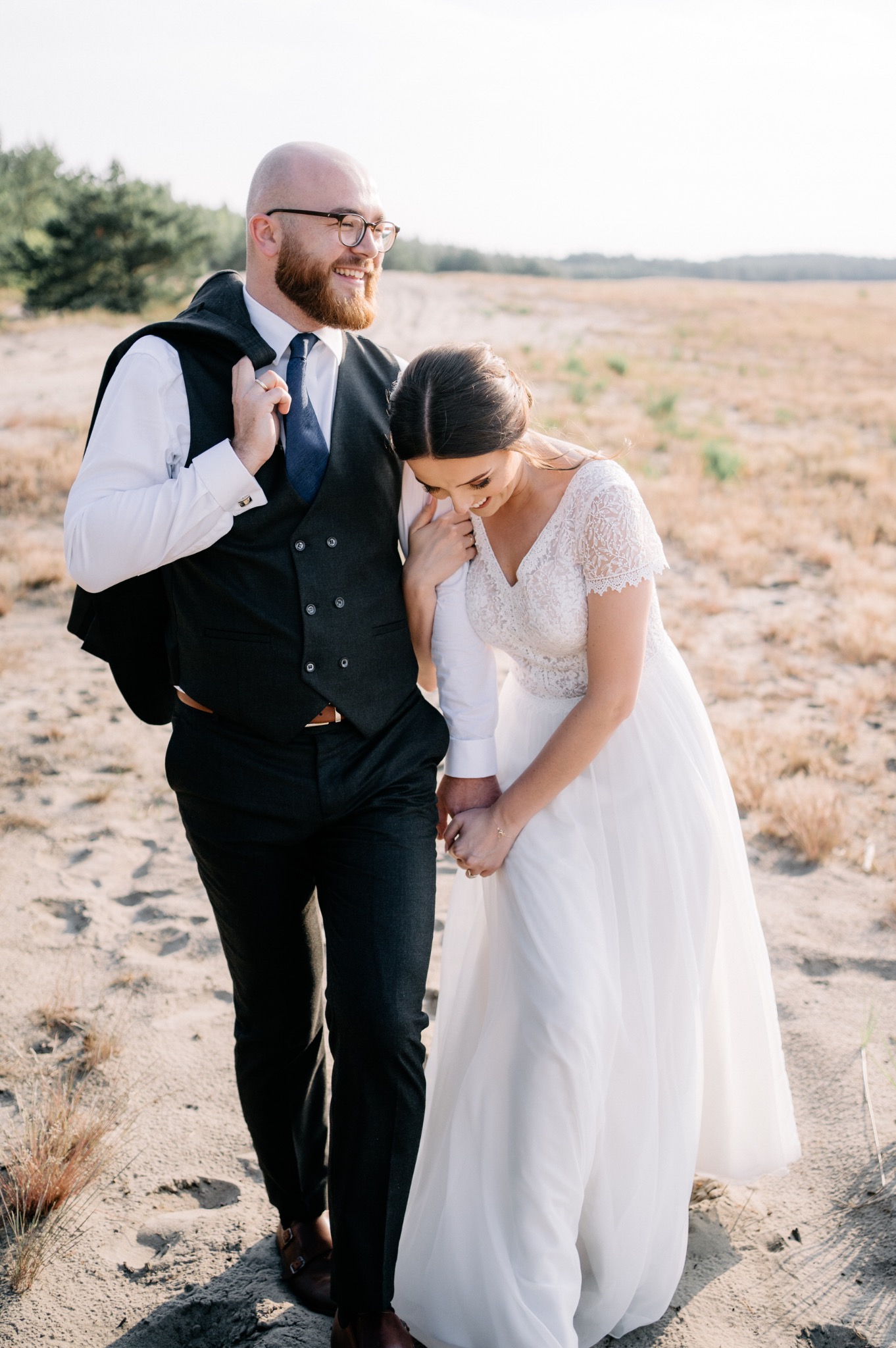 Uśmiechnięta para młoda w strojach ślubnych spaceruje po piaszczystym terenie, panna młoda w koronkowej sukni z delikatnym makijażem, pan młody w eleganckim garniturze z kamizelką i marynarką...