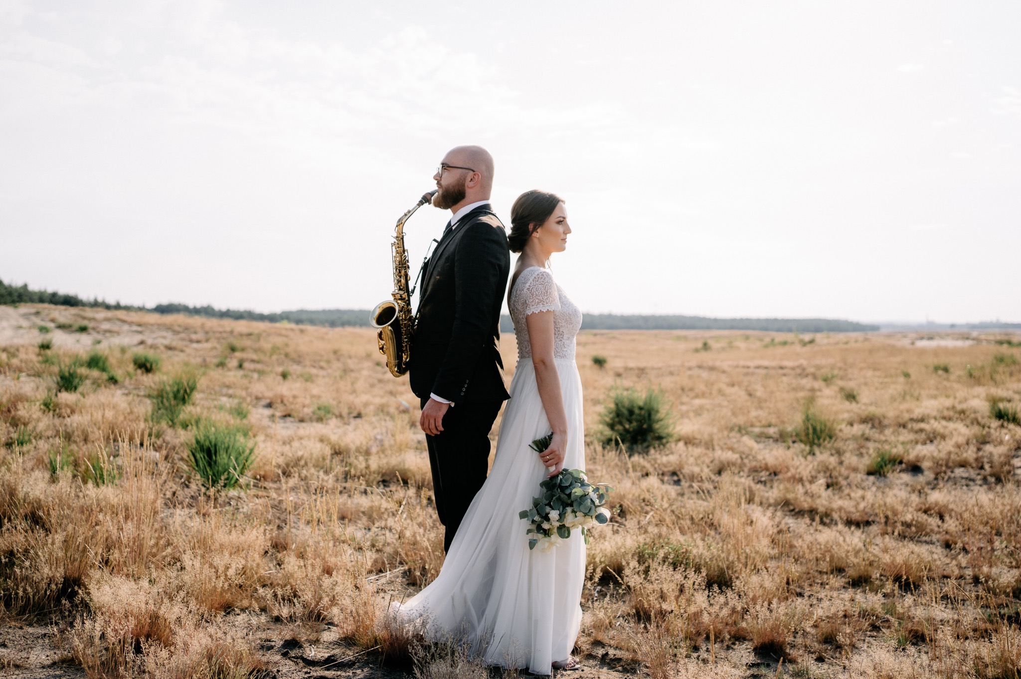 Para młoda stoi plecami do siebie na tle suchej trawy; mężczyzna trzyma saksofon.
