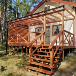 Drewniana konstrukcja domu szkieletowego z zadaszonym tarasem, schodami i balustradą w naturalnym odcieniu drewna, na tle lasu.