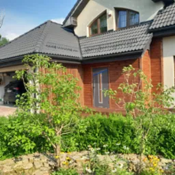 Drewniana elewacja domu jednorodzinnego z szarą stolarką okienną i drzwiową, widok na garaż z zaparkowanym samochodem, ozdobne krzewy i kwiaty w ogrodzie.
