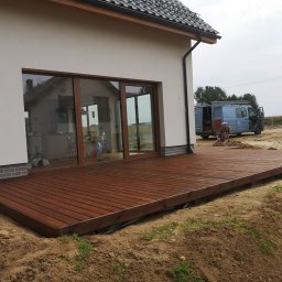 DANLUK KONSTRUKCJE DREWNIANE Łukasz Dąbrowski - Nowo wybudowany taras z brązowych desek przy białym domu z dużymi przesuwnymi drzwiami; w tle zaparkowany bus i pracownik wyrównujący teren.