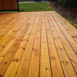 Świeżo zaimpregnowana, złocista powierzchnia z desek na zewnątrz budynku, z widoczną strukturą drewna i fragmentem zabudowy oraz trawnikiem w tle.