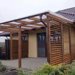 Zadaszona konstrukcja z drewnianymi lamelami przylegająca do budynku z brązową dachówką, widoczny system rynnowy i fragment ogrodu z roślinnością.