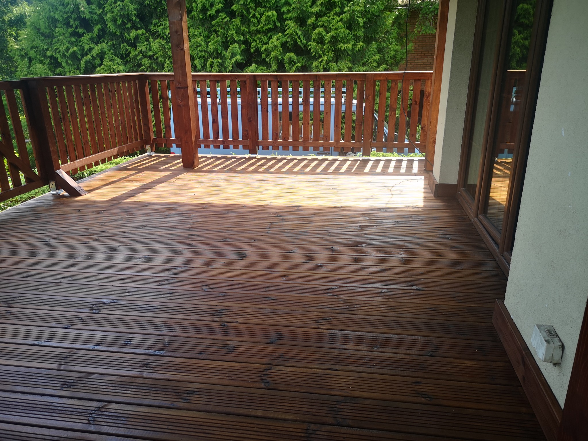 Błyszczący, drewniany taras z brązową balustradą i słupami, przylegający do budynku z oknami i zielonymi drzewami w tle.