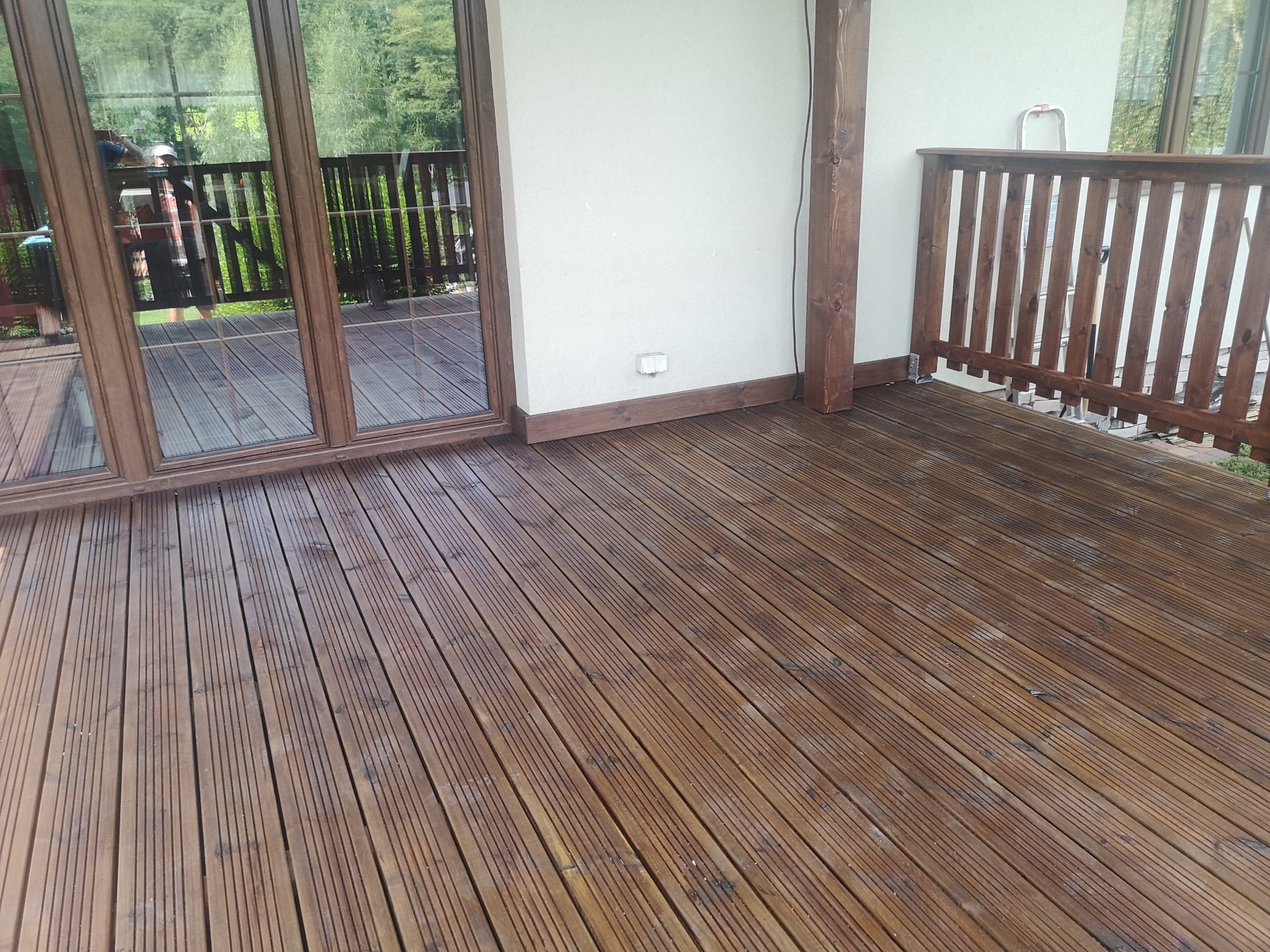 Świeżo wykończony taras z desek, brązowe balustrady i słupki, widoczne drzwi balkonowe z odbiciem osoby, biała ściana budynku z gniazdkiem elektrycznym na zewnątrz.