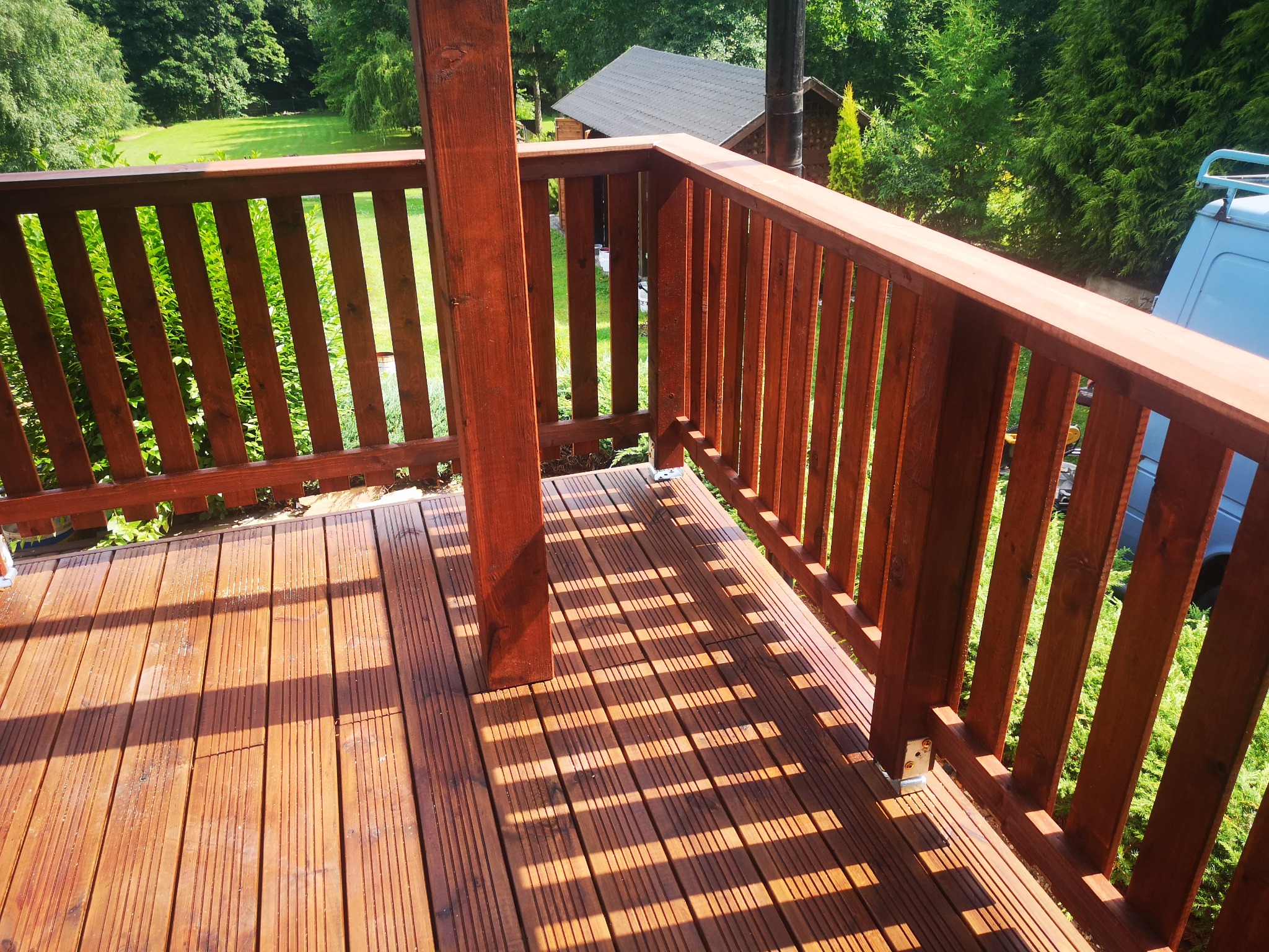 Świeżo zaimpregnowany taras z drewnianą balustradą w odcieniach brązu, z widocznymi promieniami słońca tworzącymi wzór cieni na deskach, w tle zieleń ogrodu i fragment budynku z dachem dwuspadowym.