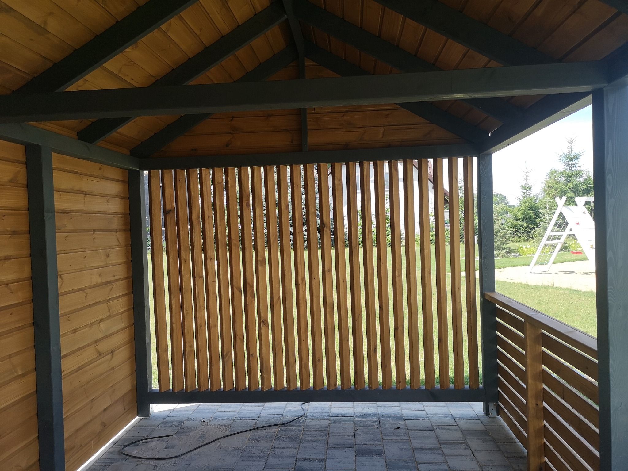 Zadaszona konstrukcja z drewnianymi ścianami z pionowych listew i drewnianą podłogą z kostki, widok na trawnik i elementy placu zabaw w tle.