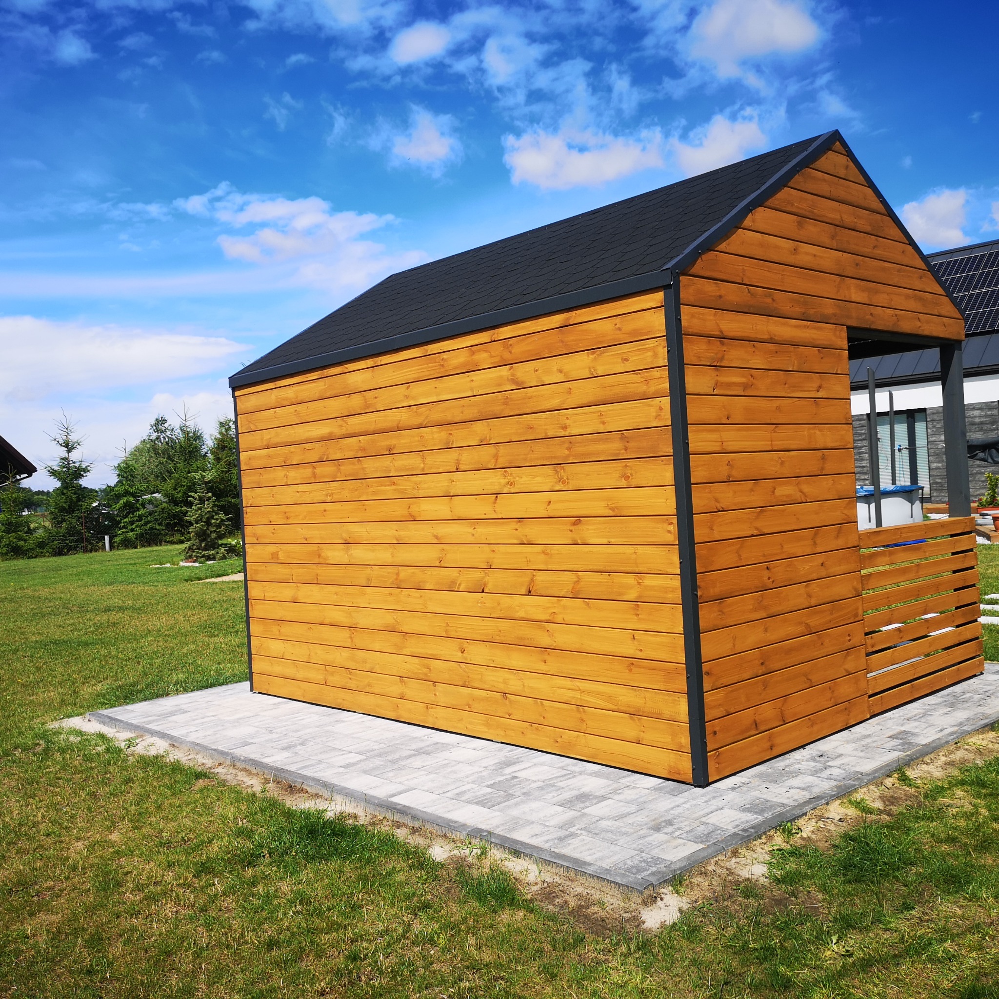 Nowoczesna, drewniana konstrukcja z czarnym dachem na kostce brukowej, otoczona trawnikiem i zielenią, widoczna w słoneczny dzień z błękitnym niebem i chmurami.
