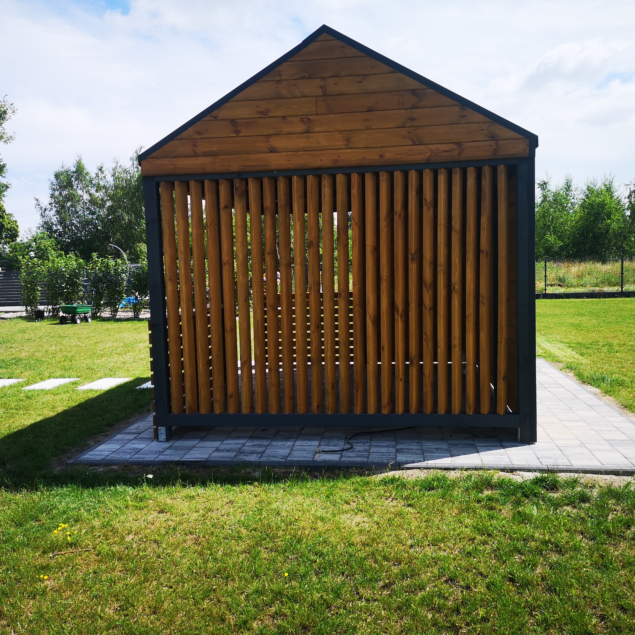 Nowoczesna, drewniana konstrukcja z pionowymi listwami, prawdopodobnie altana lub pergola, na trawniku w słoneczny dzień.