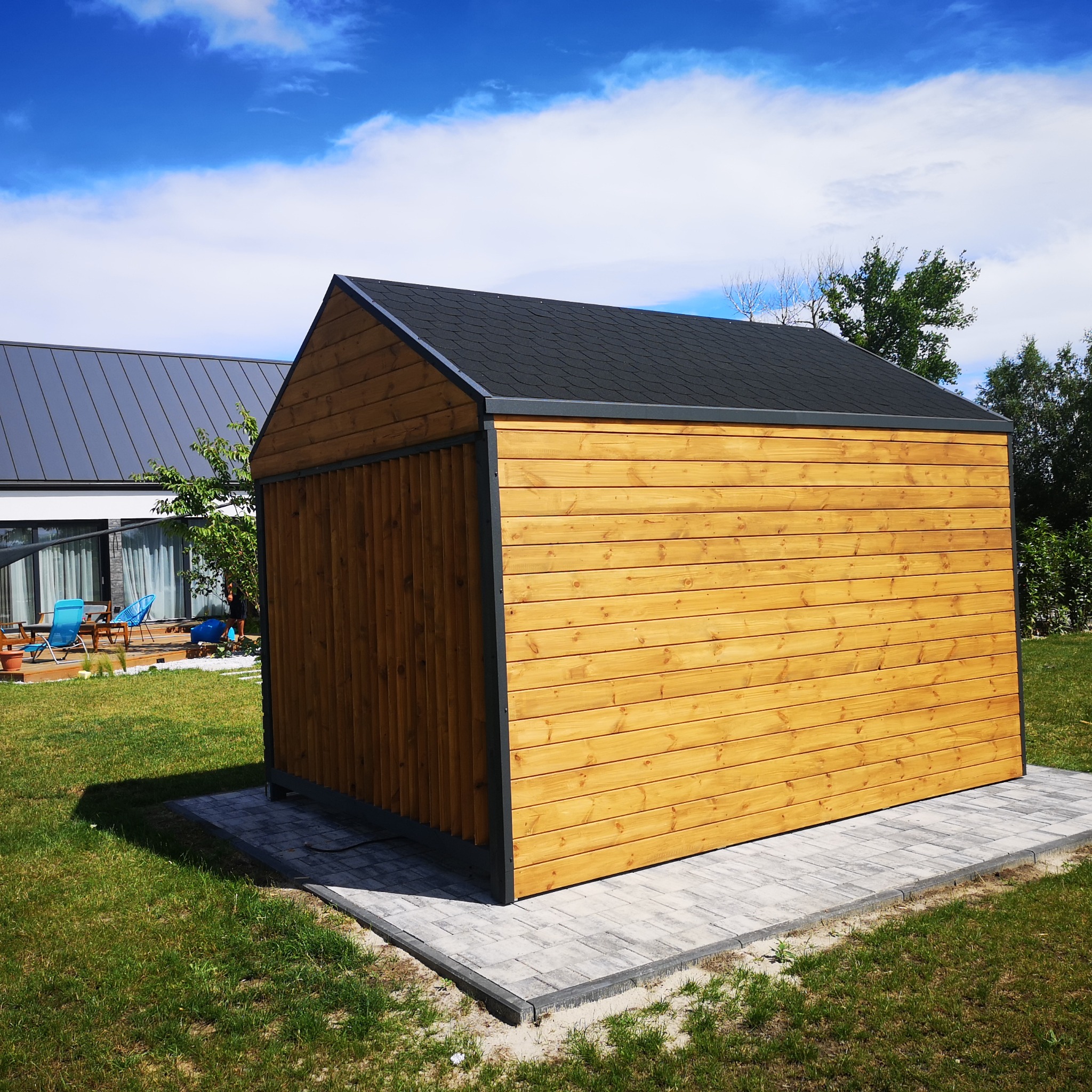 Nowoczesna, drewniana konstrukcja z czarnym dachem, stojąca na szarej kostce brukowej w ogrodzie, z widocznym domem w tle.