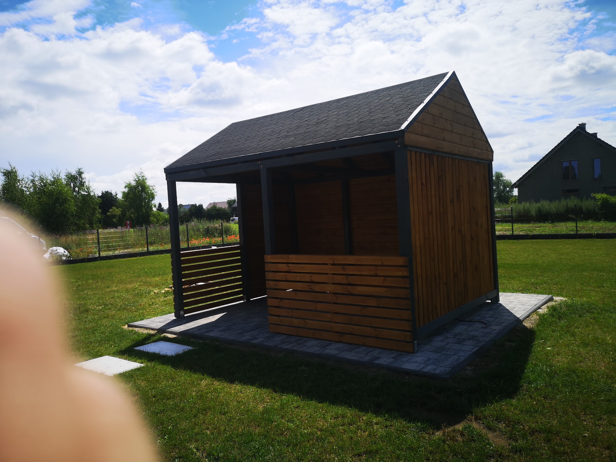 Nowoczesna altana z drewnianą zabudową i czarnym dachem bitumicznym, posadowiona na kostce brukowej w ogrodzie z widokiem na dom.