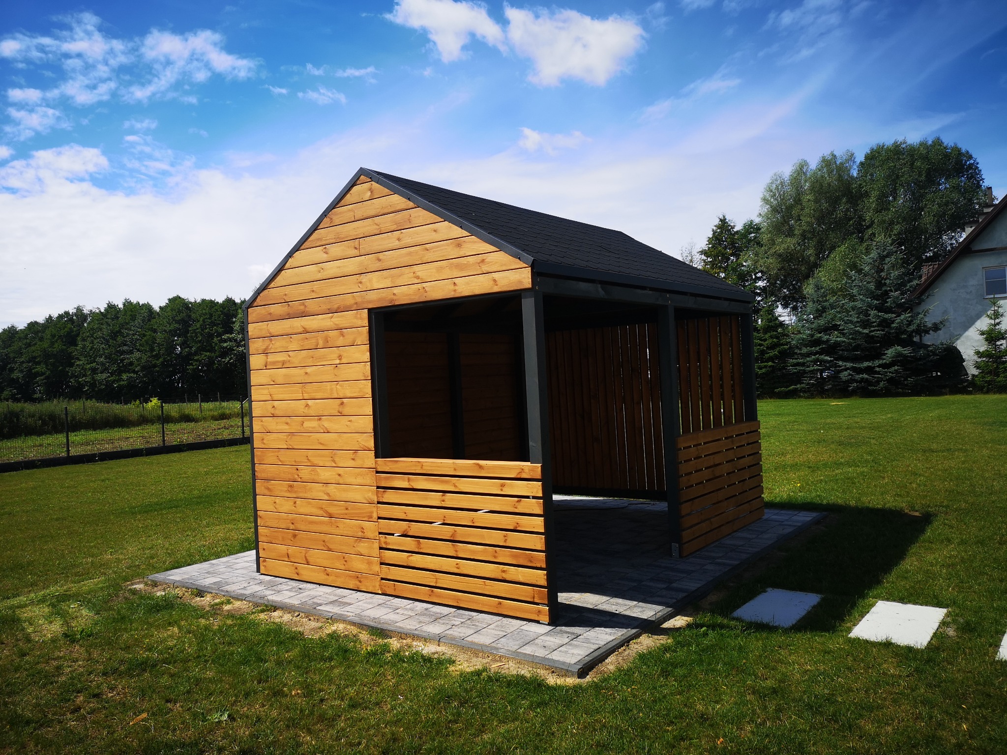 Nowoczesna altana z jasnego drewna, z czarnym dachem krytym papą, posadowiona na kostce brukowej, otoczona zielonym trawnikiem w słoneczny dzień.