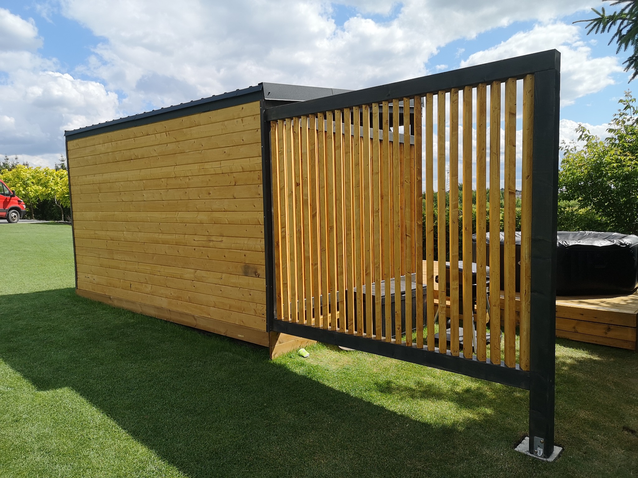 Nowoczesna konstrukcja z drewna w Starogardzie Gdańskim: połączona altana z ażurową ścianą, obok jacuzzi na podeście i równo przystrzyżony trawnik.