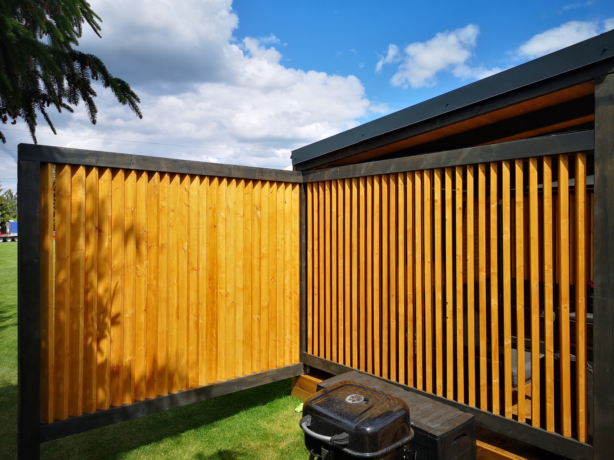 Nowoczesna konstrukcja z jasnego drewna z ciemnymi ramami, tworząca osłonę prywatności na zewnątrz, z widocznym grillem na pierwszym planie i fragmentem drzewa iglastego w górnym rogu.