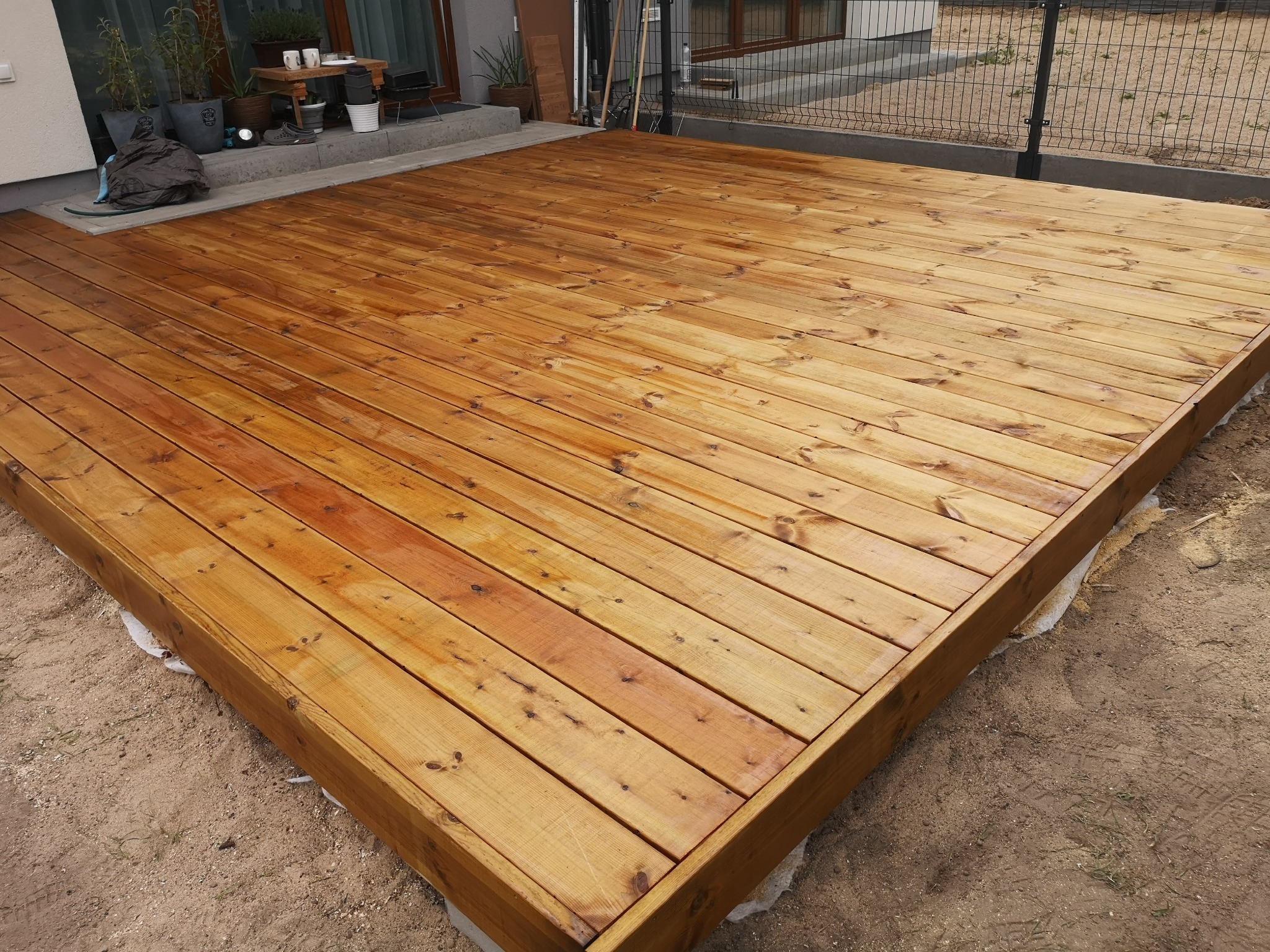 Świeżo zaimpregnowany taras z desek, widok z góry na brązowe drewno i częściowo wykończone obrzeże z widoczną folią pod spodem.