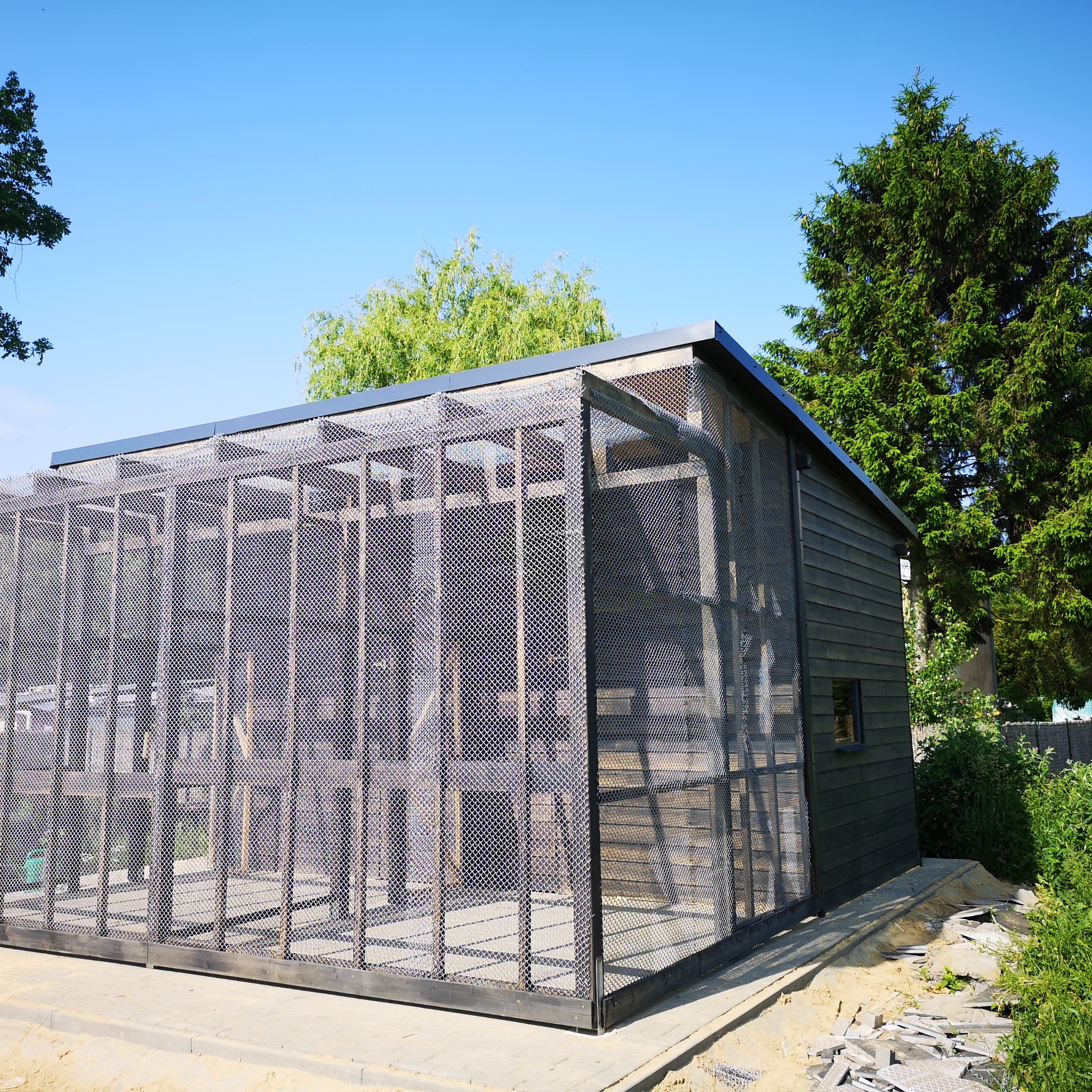 Nowoczesna, ciemna altana z drewnianą konstrukcją i metalową siatką na tle błękitnego nieba i zieleni drzew.