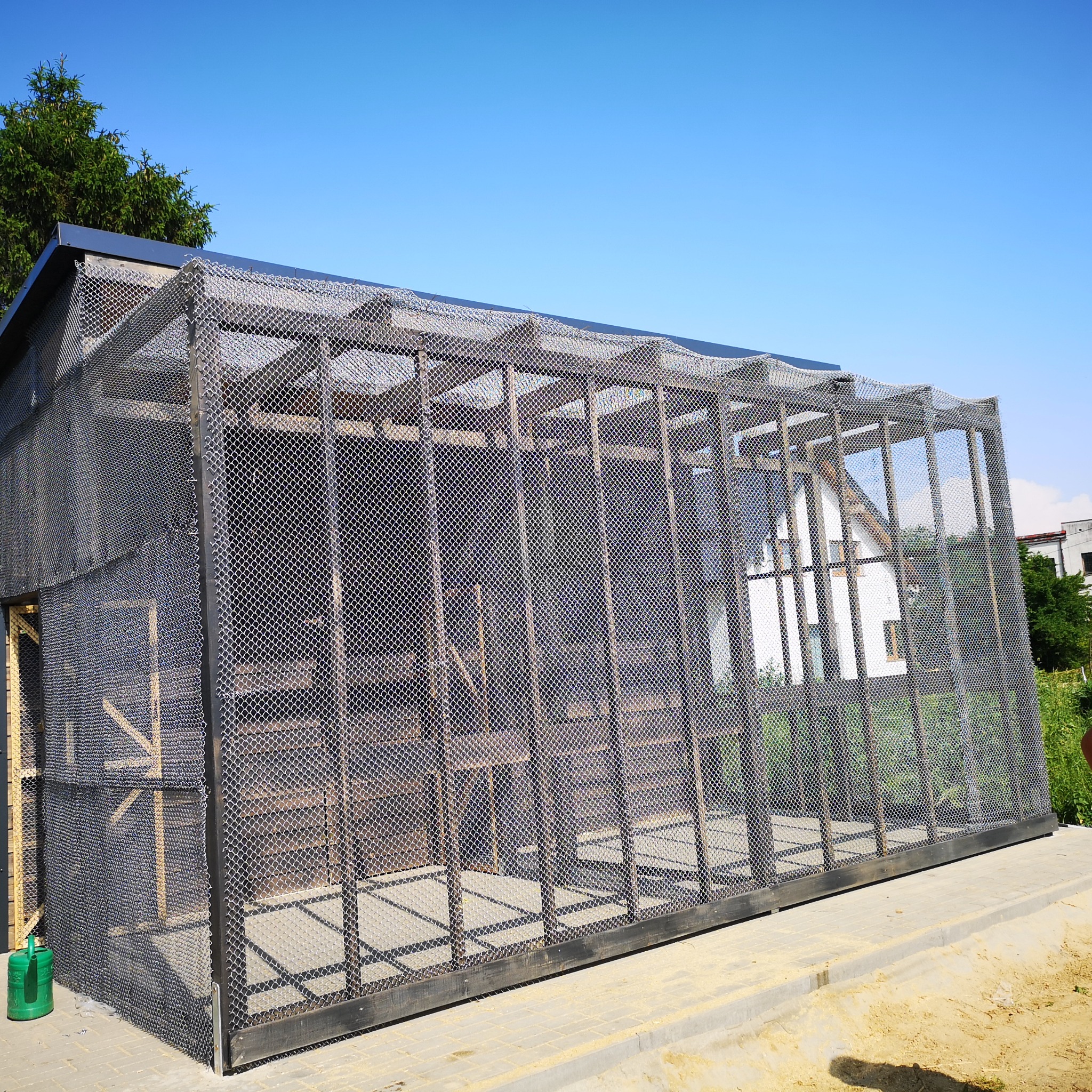 Konstrukcja szkieletowa budynku z siatką ochronną, widok z zewnątrz, z zieloną konewką przy ścianie, na tle błękitnego nieba i częściowo widocznego domu.