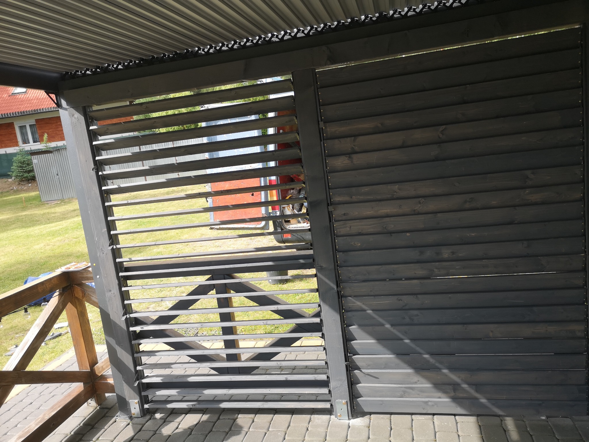 Ciemna konstrukcja z drewnianych listew, prawdopodobnie element zadaszenia tarasu lub altany, z widocznymi elementami balustrady i kostką brukową na podłożu, w tle dom z czerwonym dachem.