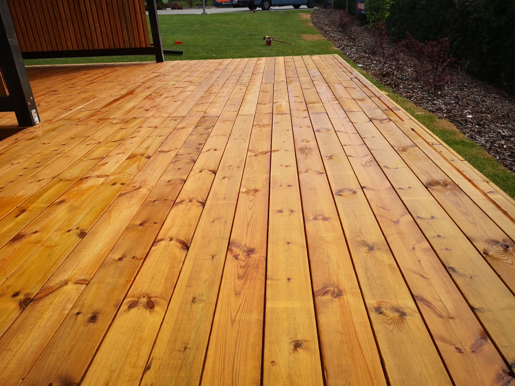 Świeżo zaimpregnowana, złocista powierzchnia z desek na zewnątrz budynku, z widoczną strukturą drewna i fragmentem zabudowy oraz trawnikiem w tle.