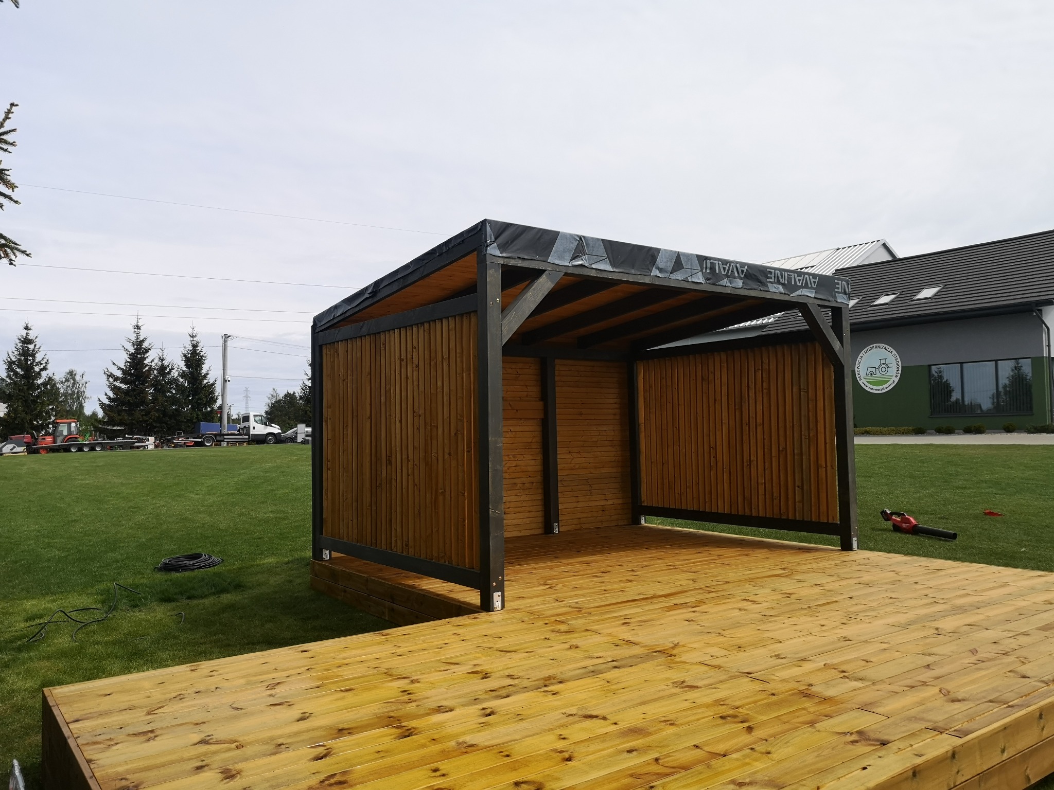 Nowoczesna konstrukcja z drewna z zadaszeniem, prawdopodobnie w trakcie budowy, widoczna na tle zielonego trawnika i budynku.