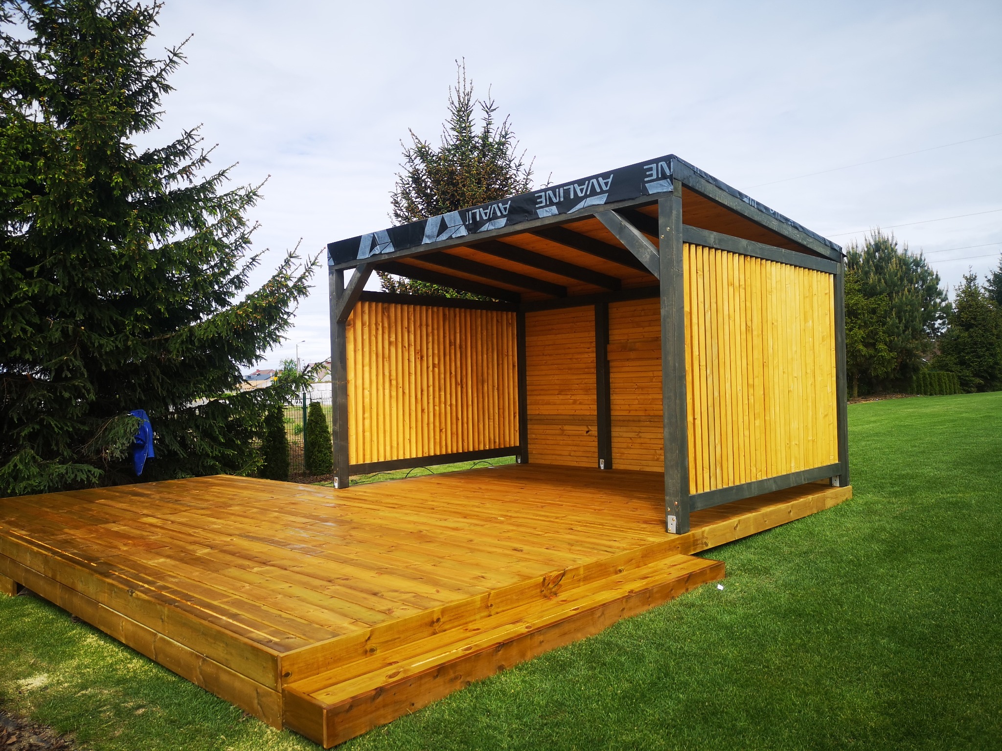 Drewniana konstrukcja z zadaszeniem na tarasie, z elementami w kolorze naturalnego drewna i ciemnoszarymi belkami, otoczona zielonym trawnikiem i drzewami iglastymi.