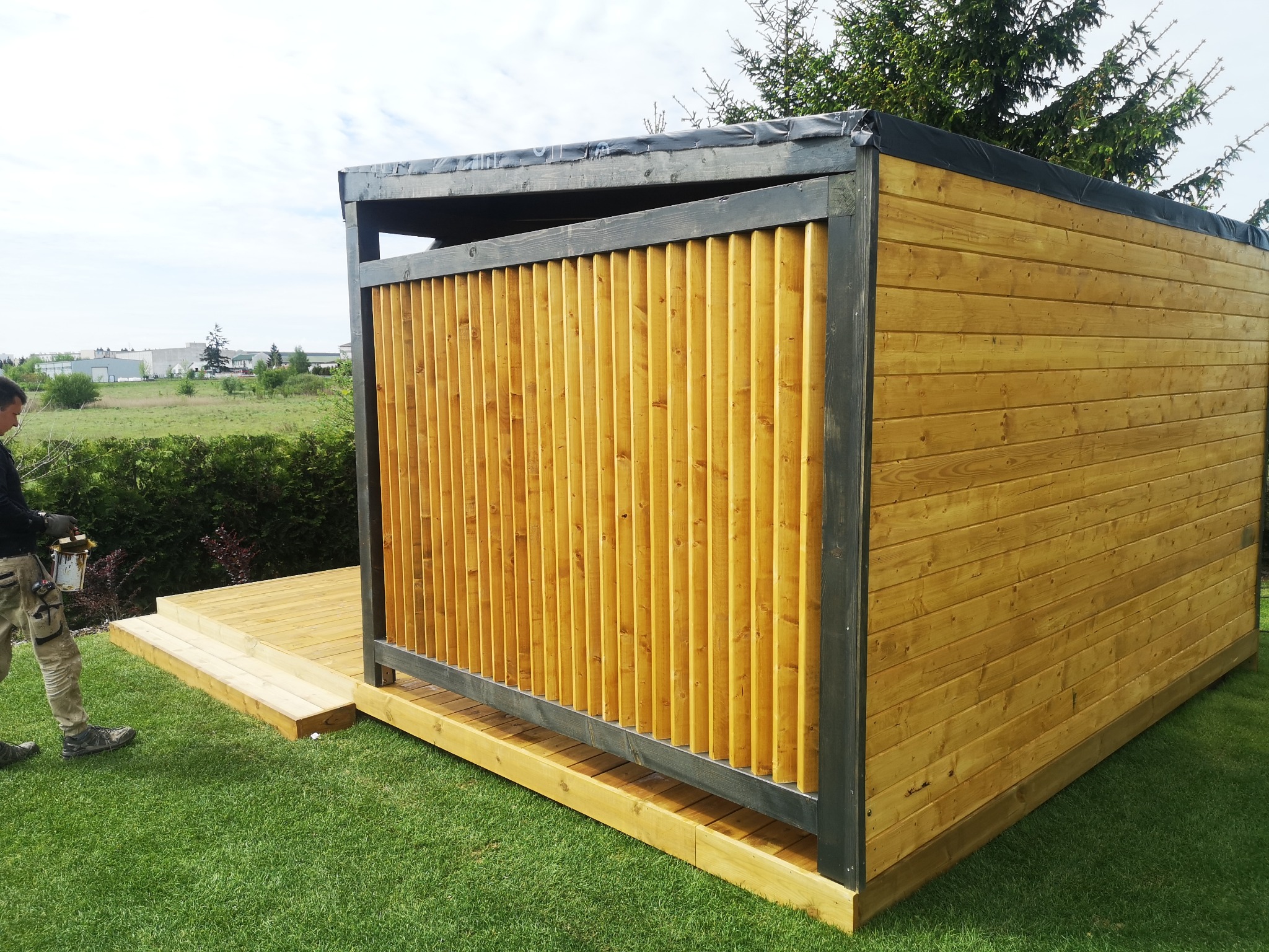 Konstrukcja z drewna, prawdopodobnie altana lub domek narzędziowy, w trakcie budowy, widoczny pracownik z puszką farby, teren zielony.