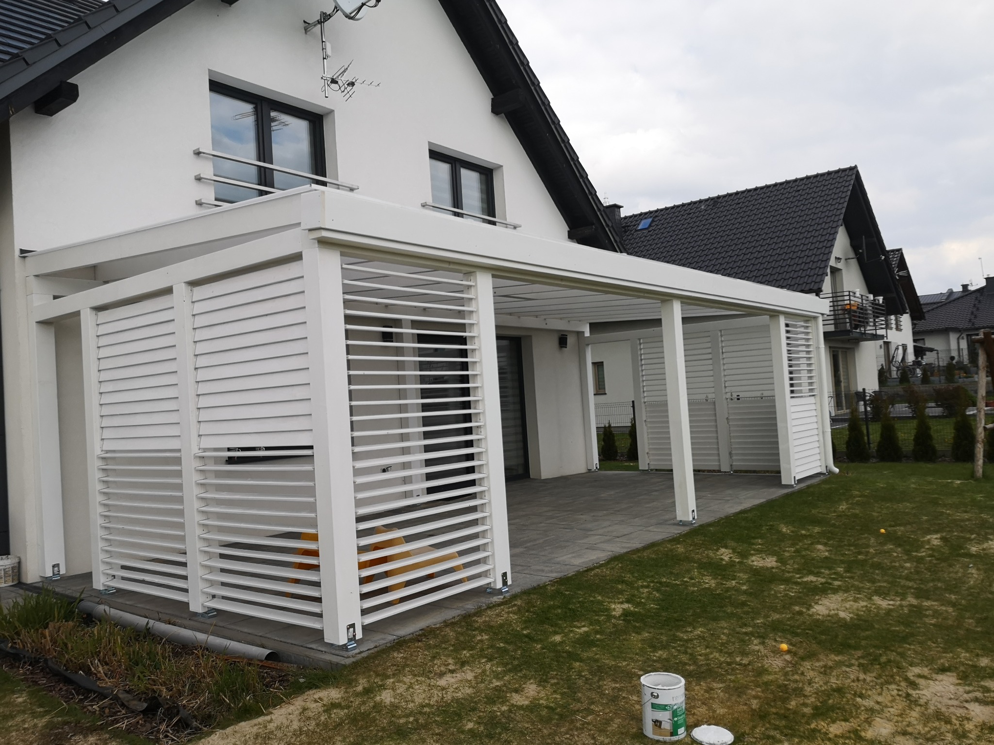 Biała konstrukcja z żaluzjowymi ścianami przylegająca do białego domu z ciemnym dachem, widoczny fragment tarasu z szarej kostki i trawnik, w tle inne domy.