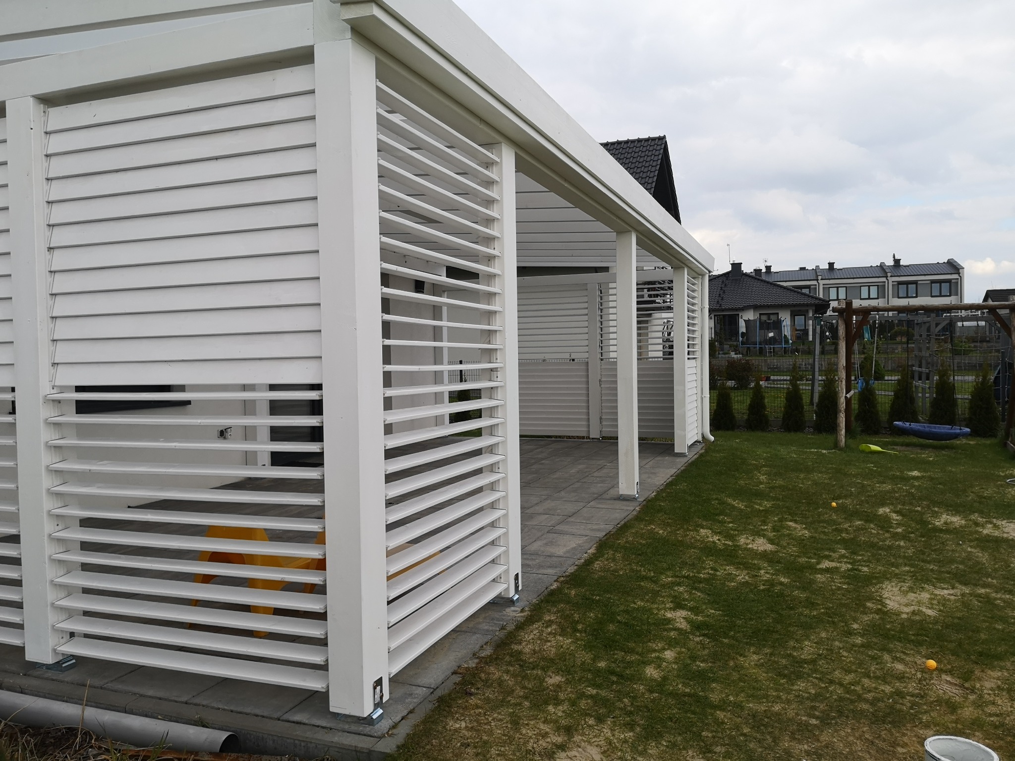 Biała konstrukcja z poziomych listew, tworząca zacienione miejsce w ogrodzie, z widocznym żółtym krzesłem wewnątrz, na tle trawnika i domów.