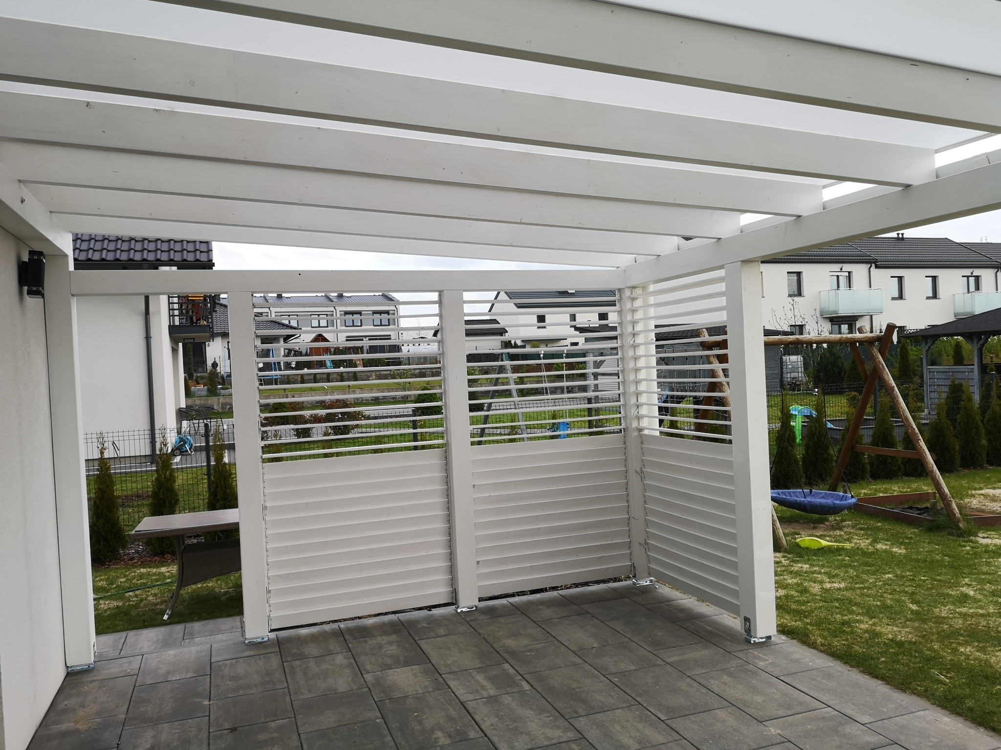 Biała pergola z drewnianymi lamelami tworząca osłonę, widoczna z zewnątrz na tle domów i trawnika z huśtawką.