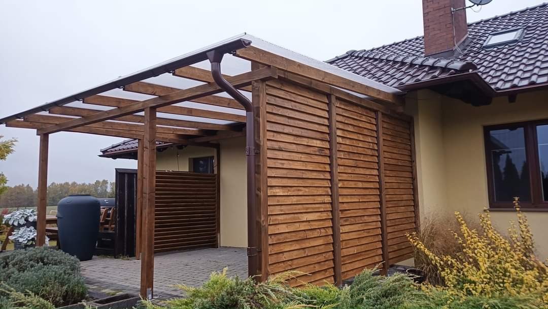 Zadaszona konstrukcja drewniana z transparentnym dachem, przylegająca do domu z brązową rynną, drewnianą ścianą z poziomych desek i brukowanym podłożem. W tle widoczny zbiornik na deszczówkę i zieleń.