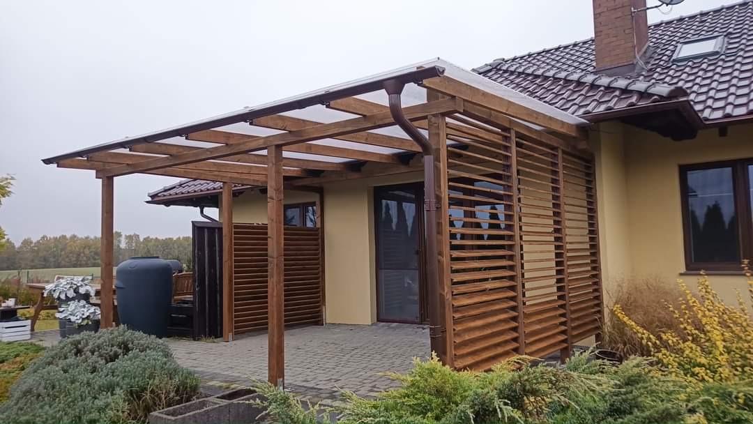 Zadaszona konstrukcja z drewnianymi lamelami przylegająca do budynku z brązową dachówką, widoczny system rynnowy i fragment ogrodu z roślinnością.