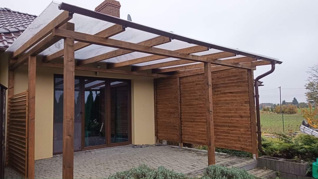 Zadaszona konstrukcja z drewna przy budynku, z częściowo zabudowanymi ścianami z desek, zadaszenie z przezroczystych paneli, widok na ogród w tle.