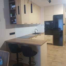 Twoje M. Meble na wymiar - Jasna kuchnia z drewnianymi blatami, czarnymi hokerami i minimalistycznymi lampami wiszącymi, wyposażona w zabudowany sprzęt AGD.