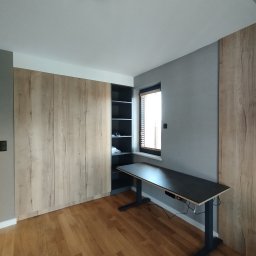 Twoje M. Meble na wymiar - Wnętrze pokoju z dużą szafą przesuwną z drewnopodobnymi frontami, regałem z czarnymi półkami, biurkiem z regulowaną wysokością blatu i drewnianą podłogą.