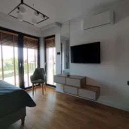 Nowoczesna sypialnia z drewnianą podłogą, łóżkiem z szarą pościelą, krzesłem obok okna z żaluzjami, wiszącą szafką RTV pod telewizorem i designerską lampą sufitową.