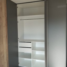Twoje M. Meble na wymiar - Otwarta szafa wnękowa z szarymi drzwiami i białym wnętrzem, widoczne półki, szuflady i drążek na ubrania, zarys podłogi zabezpieczony kartonem.