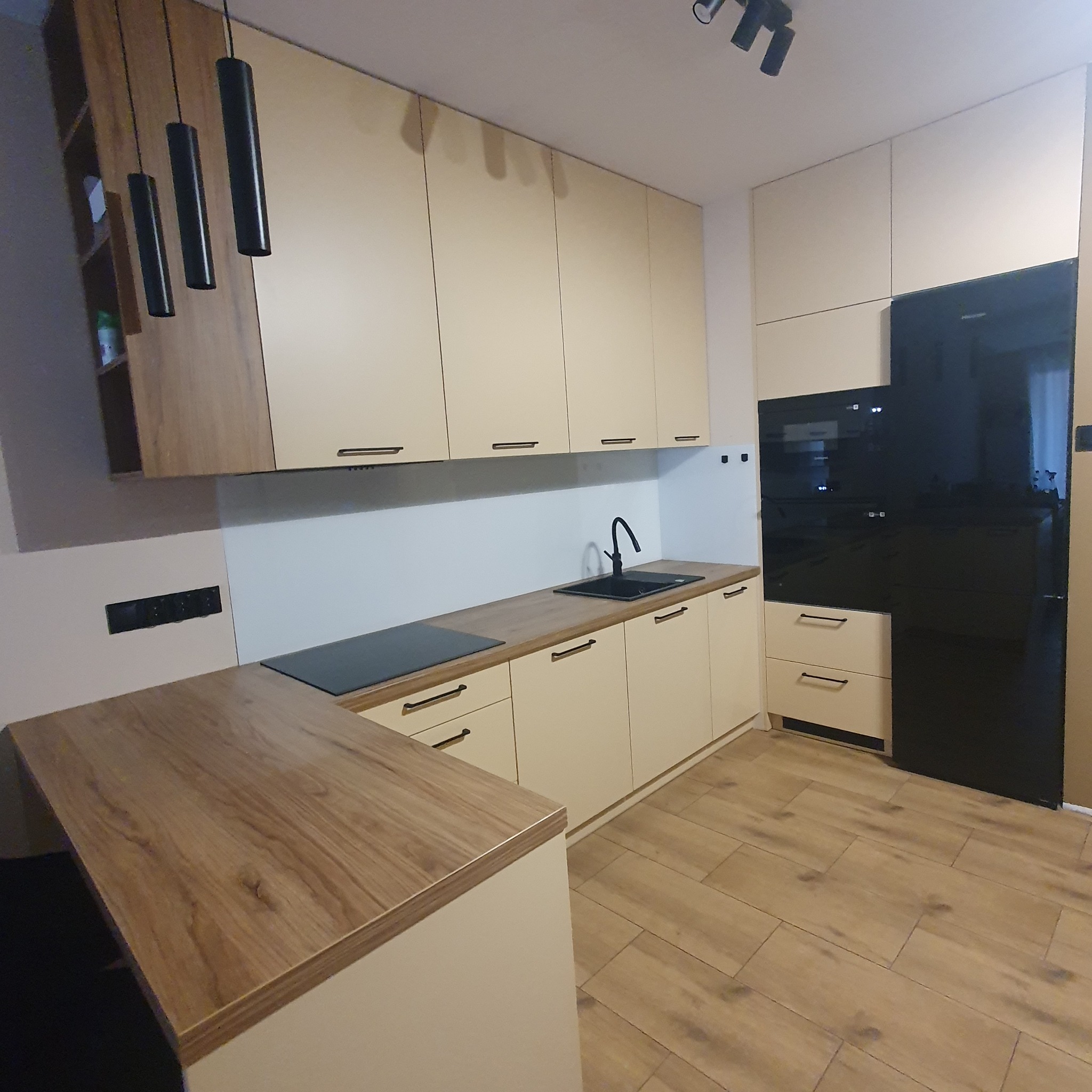 Jasna kuchnia z drewnianym blatem, czarną armaturą i oświetleniem, widok na zabudowę piekarnika i lodówki.