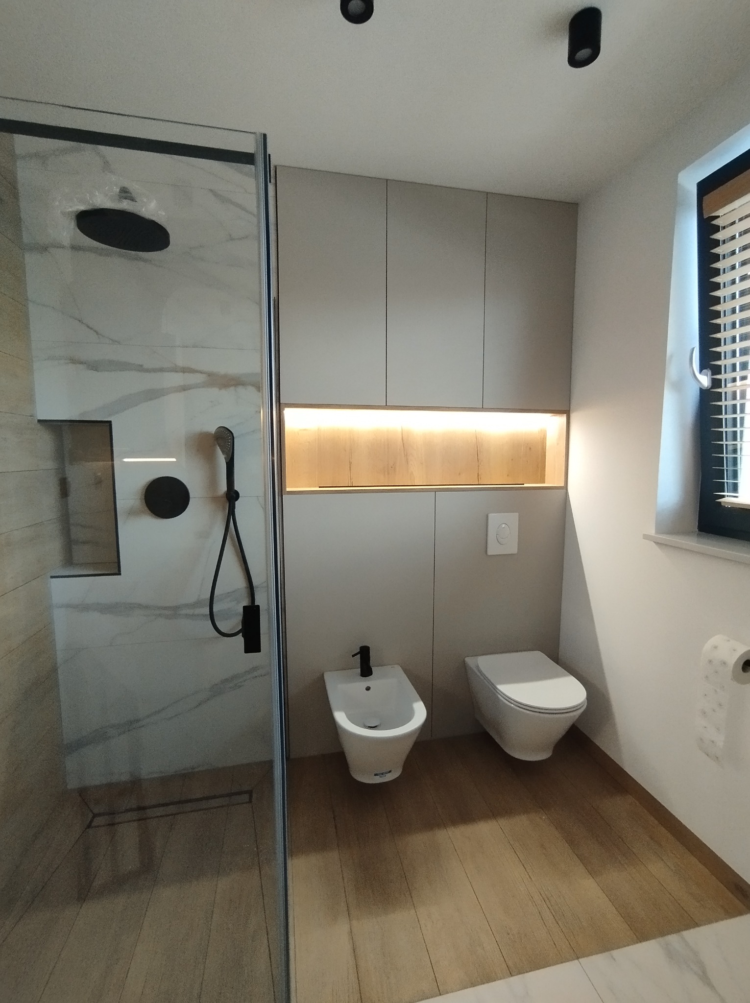 Nowoczesna łazienka z kabiną prysznicową typu walk-in wykończoną marmurem, wiszącą toaletą i bidetem, podświetlana wnęka nad zabudową sanitarną, drewnopodobna podłoga.