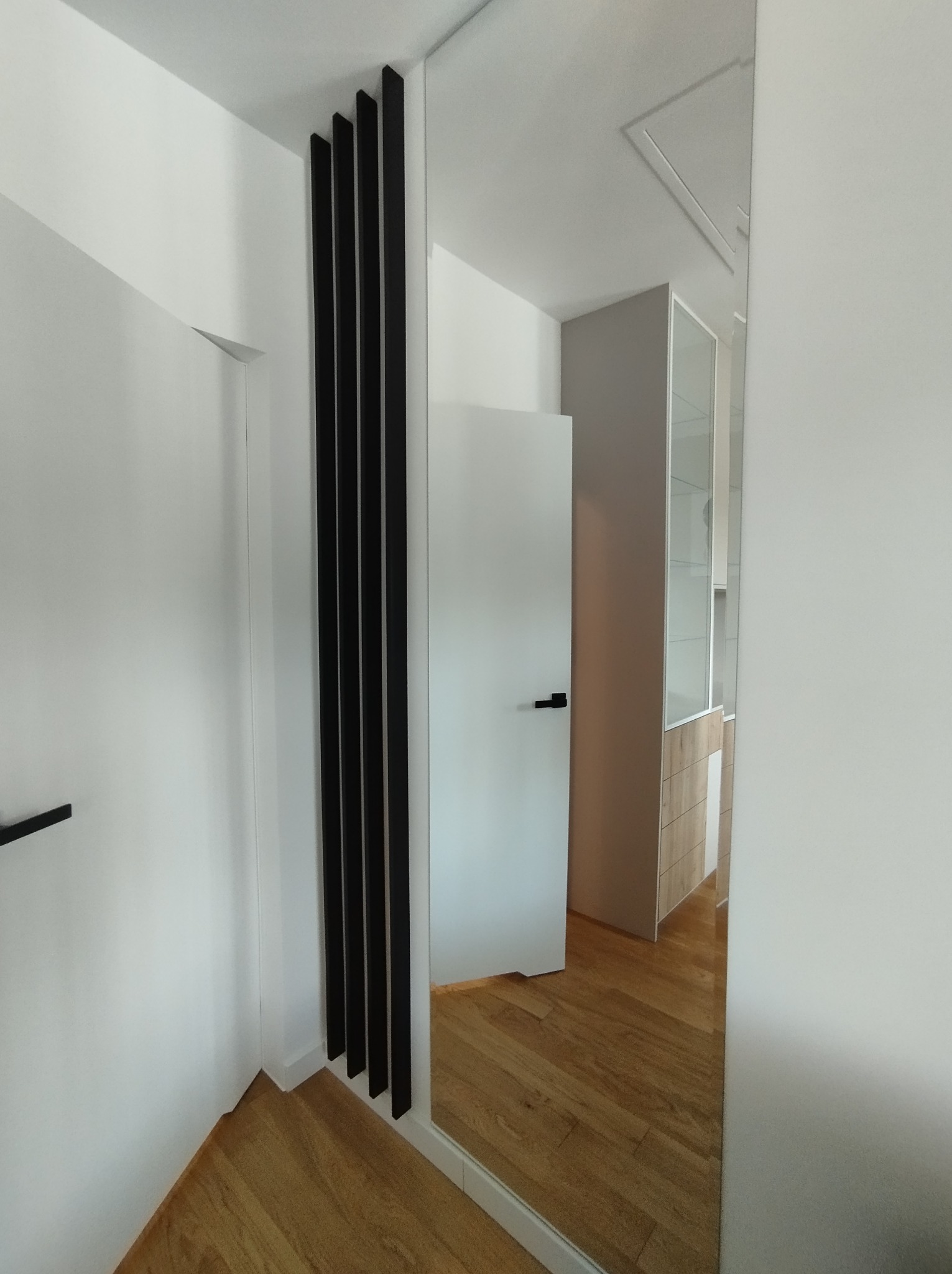 Nowoczesny przedpokój z lustrem, drewnianą podłogą i czarnymi, pionowymi listwami dekoracyjnymi na białej ścianie, odbicie szafy w lustrze.