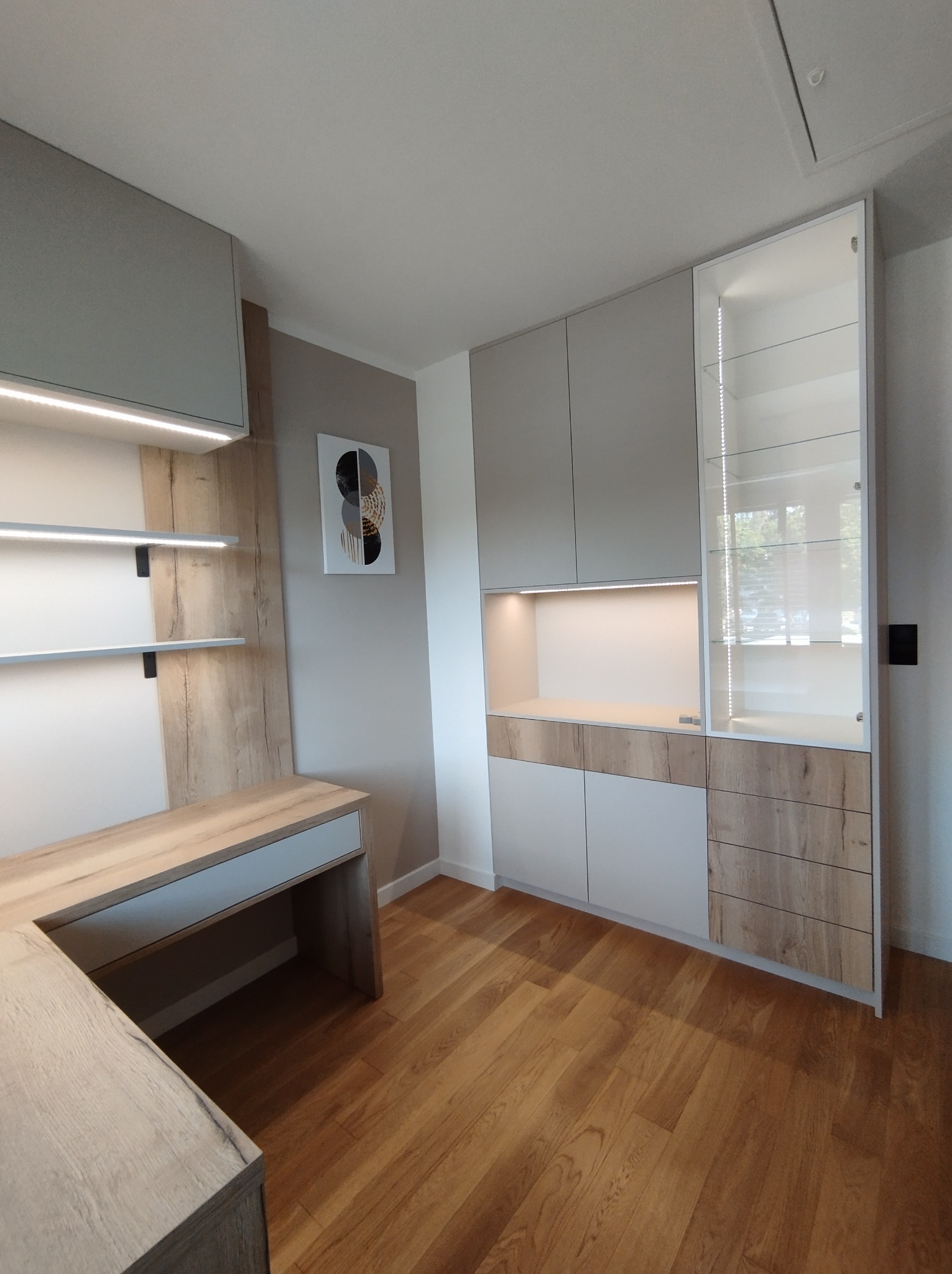 Jasna zabudowa meblowa z biurkiem w Łopienniku Dolnym-Kolonii, łącząca szafy z szufladami i przeszkloną witryną, z akcentami drewna i oświetleniem LED.