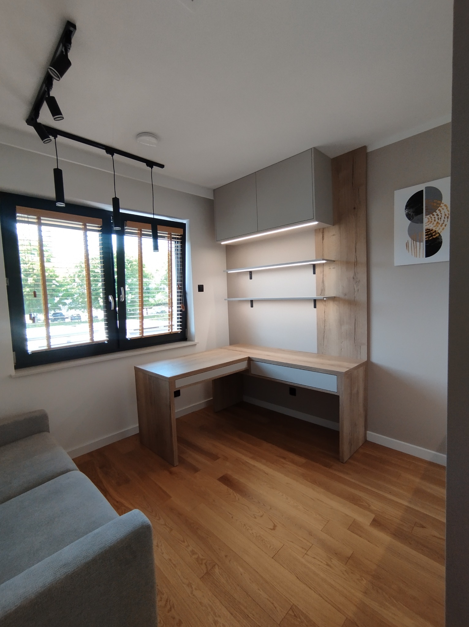 Drewniane biurko narożne z szufladami i półkami podświetlonymi LED, zamontowane przy oknie z drewnianymi żaluzjami w nowoczesnym pokoju z drewnianą podłogą i szarą sofą.