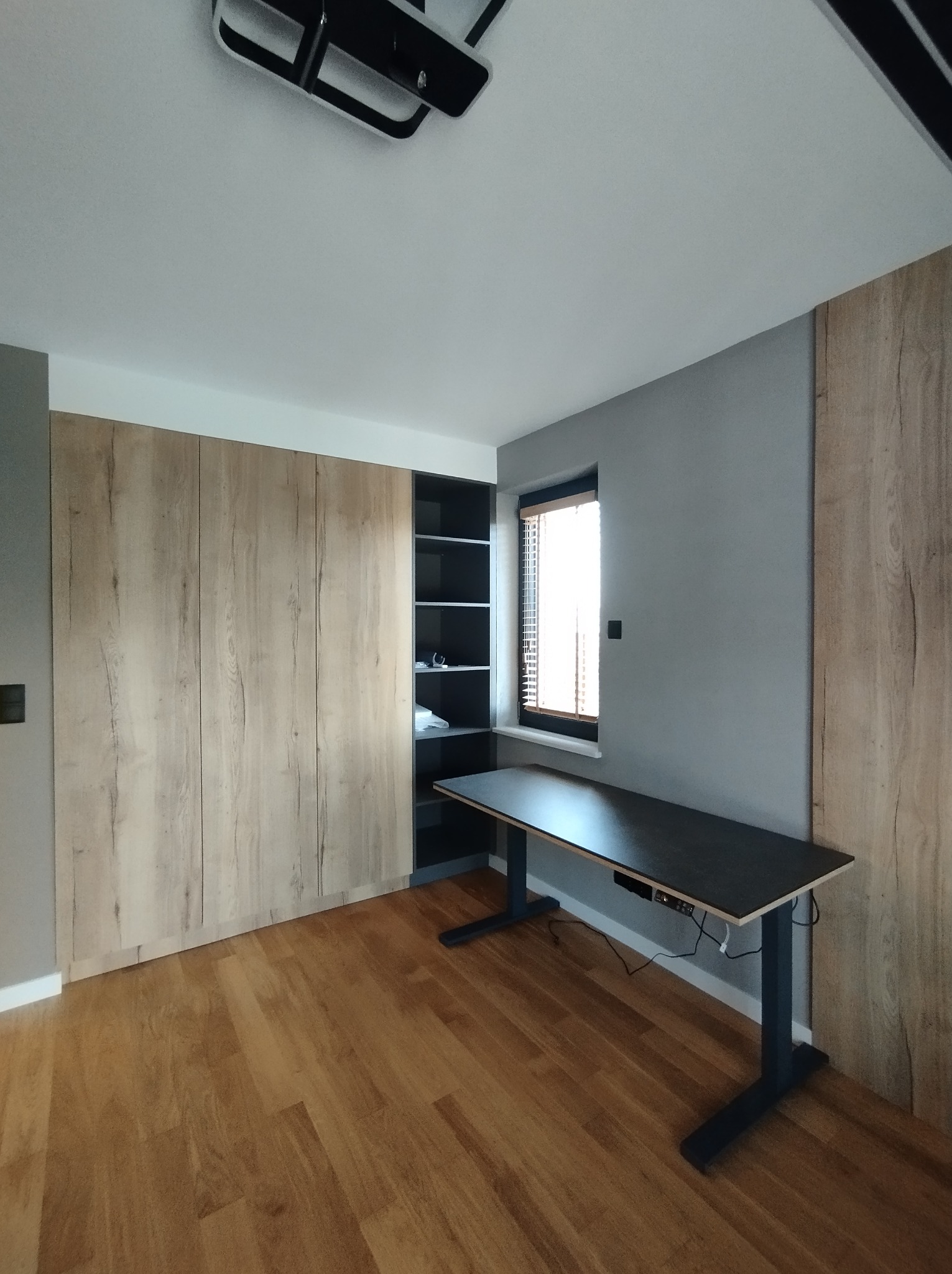 Wnętrze pokoju z dużą szafą przesuwną z drewnopodobnymi frontami, regałem z czarnymi półkami, biurkiem z regulowaną wysokością blatu i drewnianą podłogą.